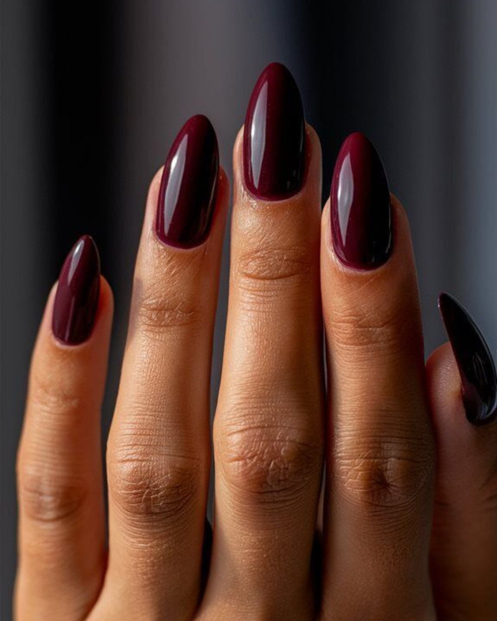Close-up of long almond-shaped nails painted a glossy deep burgundy against a soft dark grey background