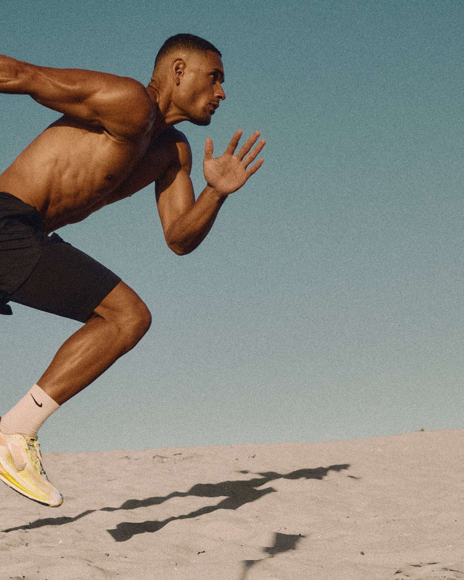 Shirtless runner in black shorts and yellow sneakers sprinting up a sand dune under a blue sky