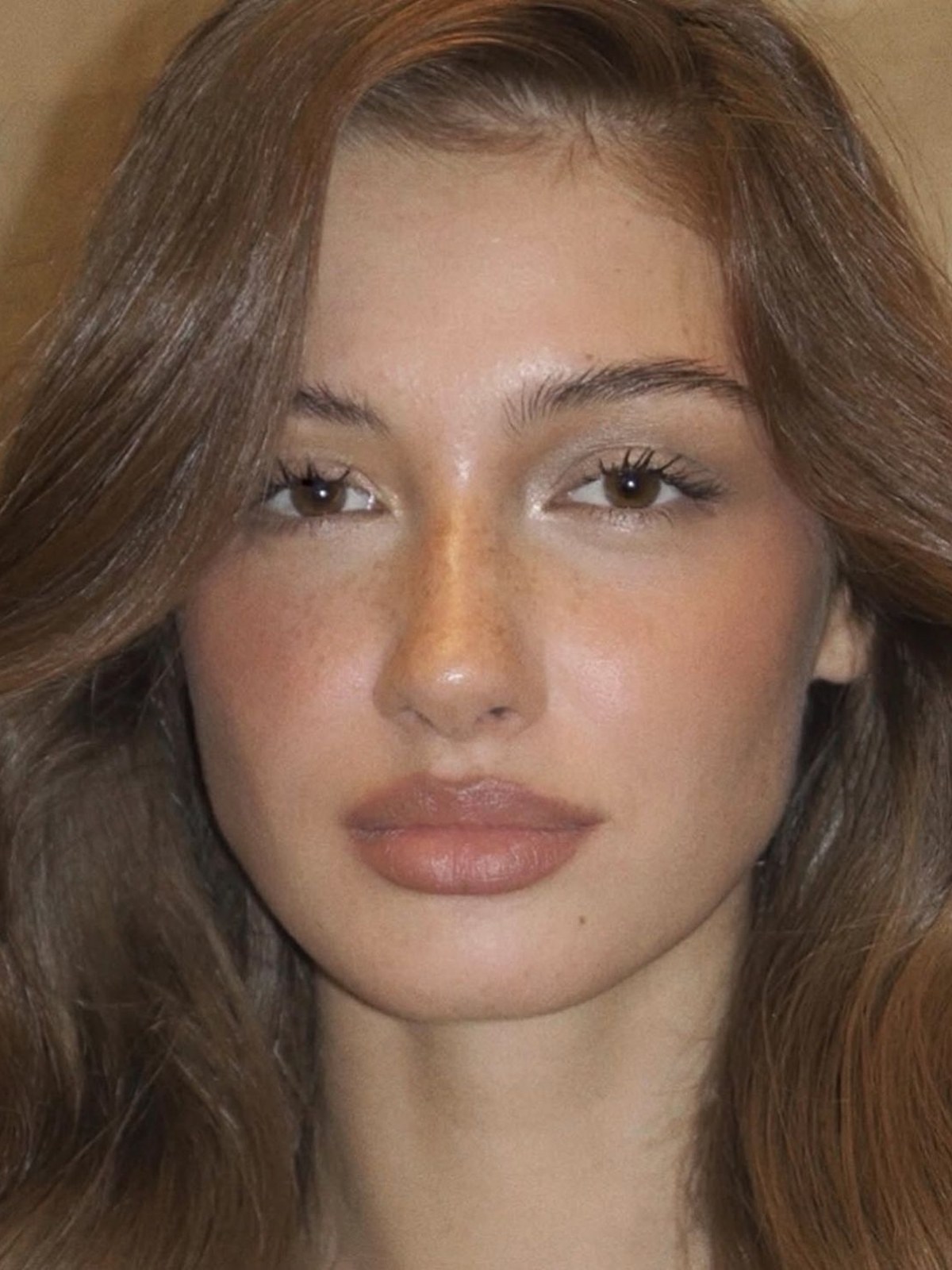 Woman with wavy auburn hair, soft bronzed makeup, a glowing nose and a nude brown lip, against a warm beige backdrop