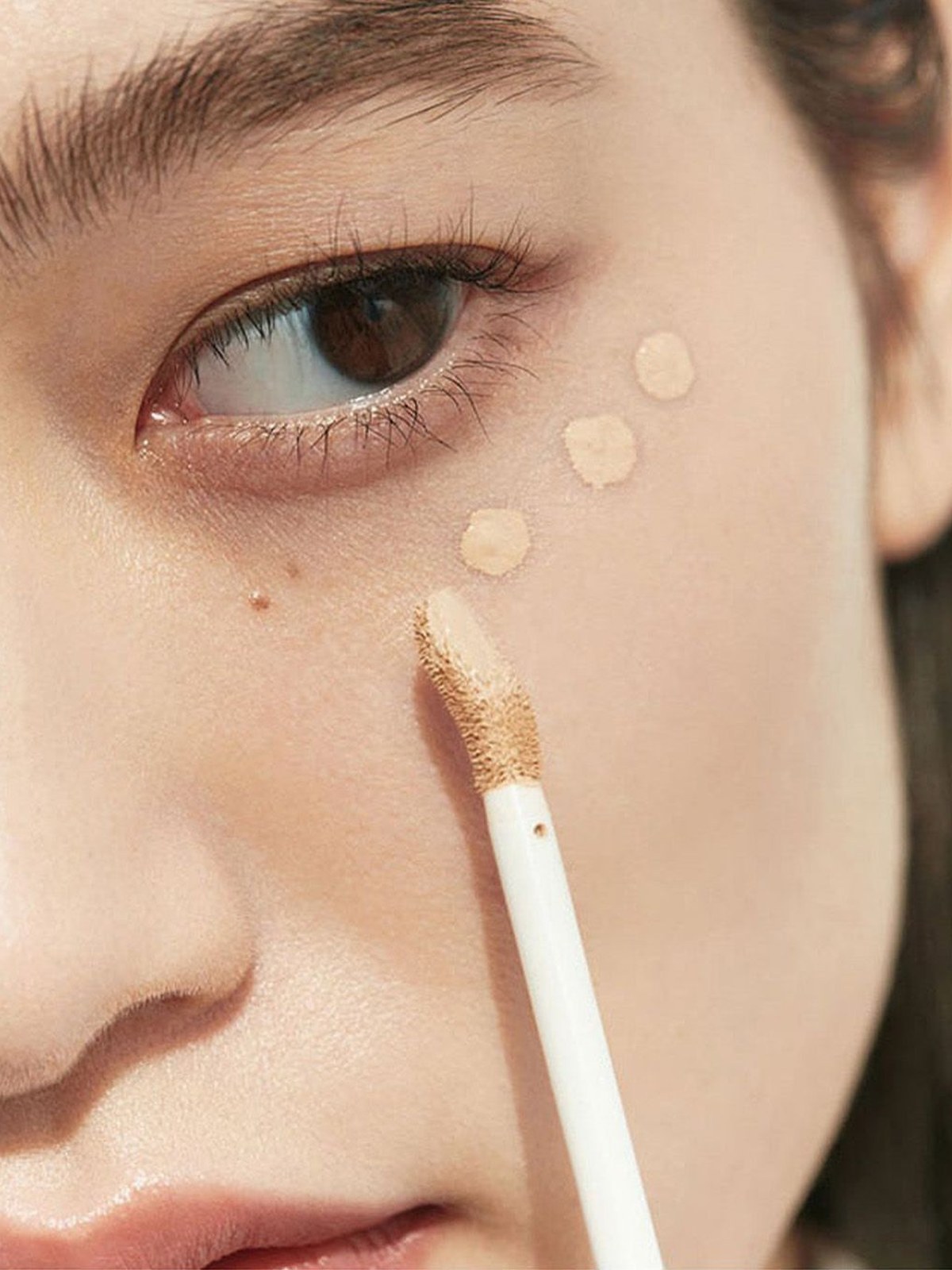 Close-up of a woman with a straight dark fringe, a slicked-back ponytail and dots of concealer on her cheek, holding a white concealer applicator