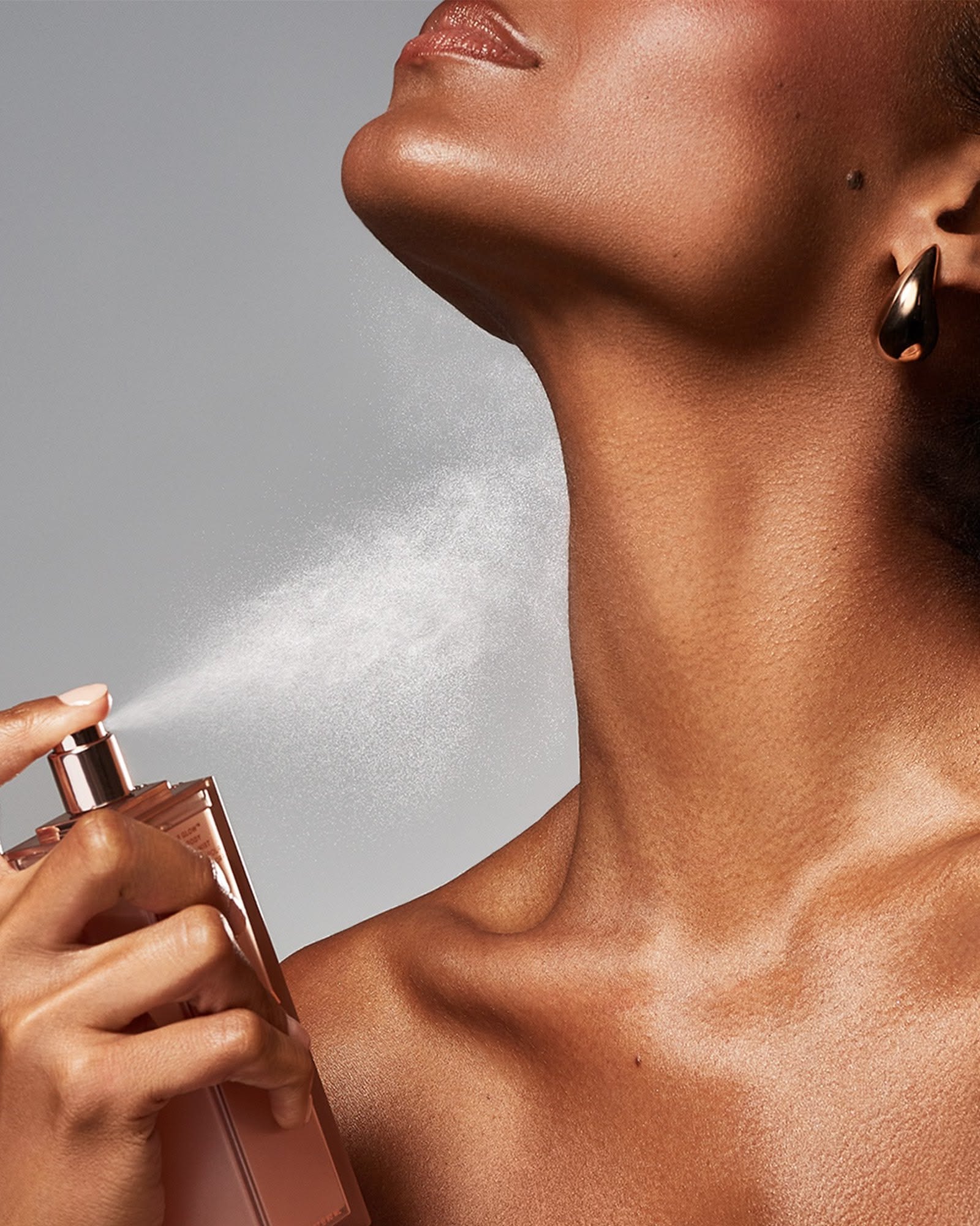 Close up of a woman's neck and shoulder as a perfume mist is sprayed from a rose gold bottle against a grey background