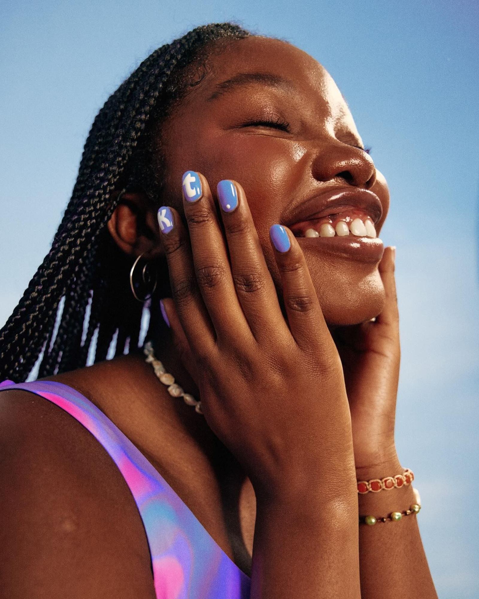 A woman with long box braids laughs with her eyes closed and both hands at her cheek, showing periwinkle blue nails with white letters, a pearl necklace, bracelets and a pink and purple top under a blue sky.