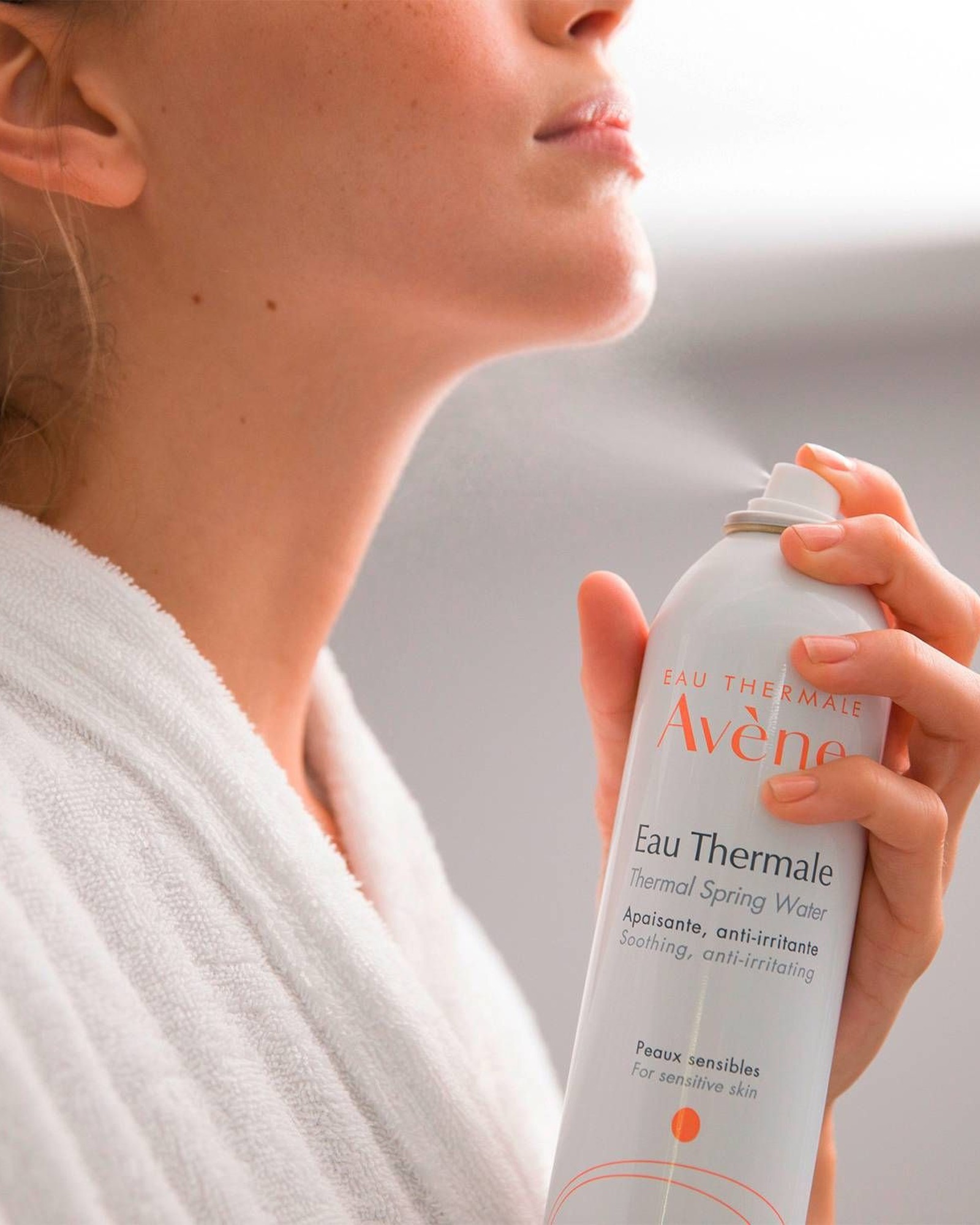 Close-up of a woman in a white terry robe holding a can of Eau Thermale Avène Thermal Spring Water and spraying her neck and chin