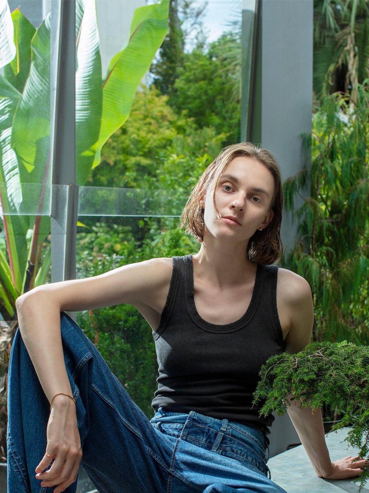 Model seated in a black tank and blue jeans beside a greenhouse window