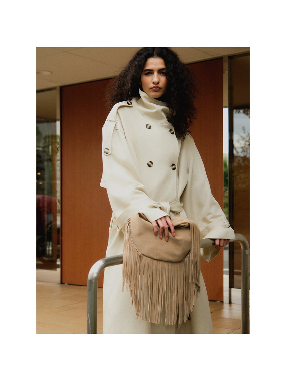 Model seated in a white coat with a fringed tan handbag
