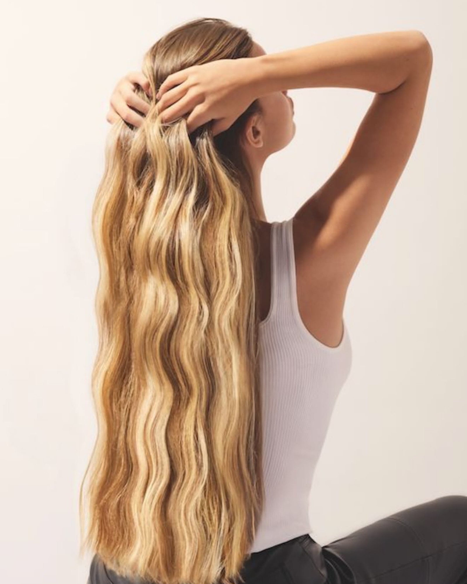 Woman seen from behind with long wavy golden blonde hair, arms raised into her hair, in a white ribbed tank top
