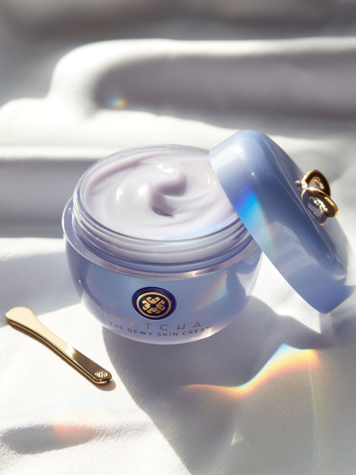 Open jar of Tatcha The Dewy Skin Cream with a gold spatula on white fabric in soft light with rainbow reflections
