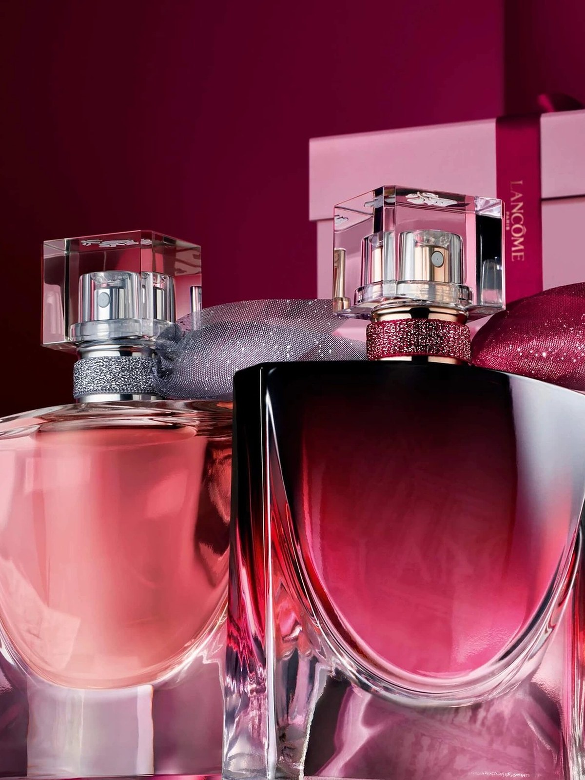 Two La Vie Est Belle perfume bottles against a deep burgundy backdrop