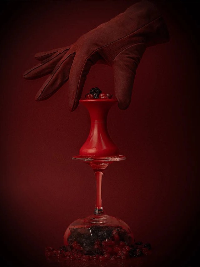 A dark gloved hand holds a red bottle above a glass and berries.