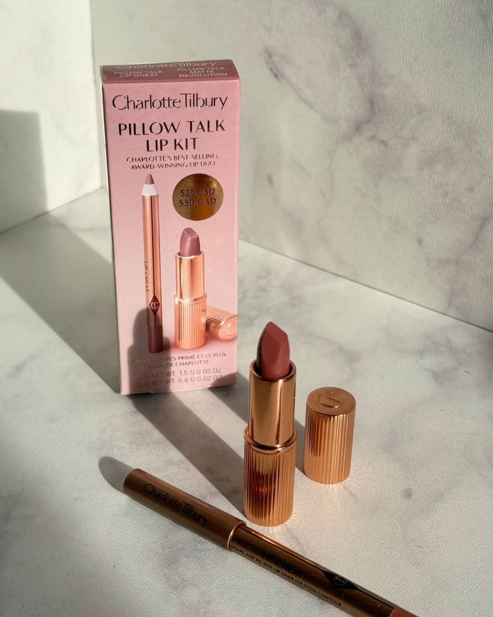 Rose-gold lipstick and lip pencil beside a pink Pillow Talk Lip Kit box on a sunlit marble surface