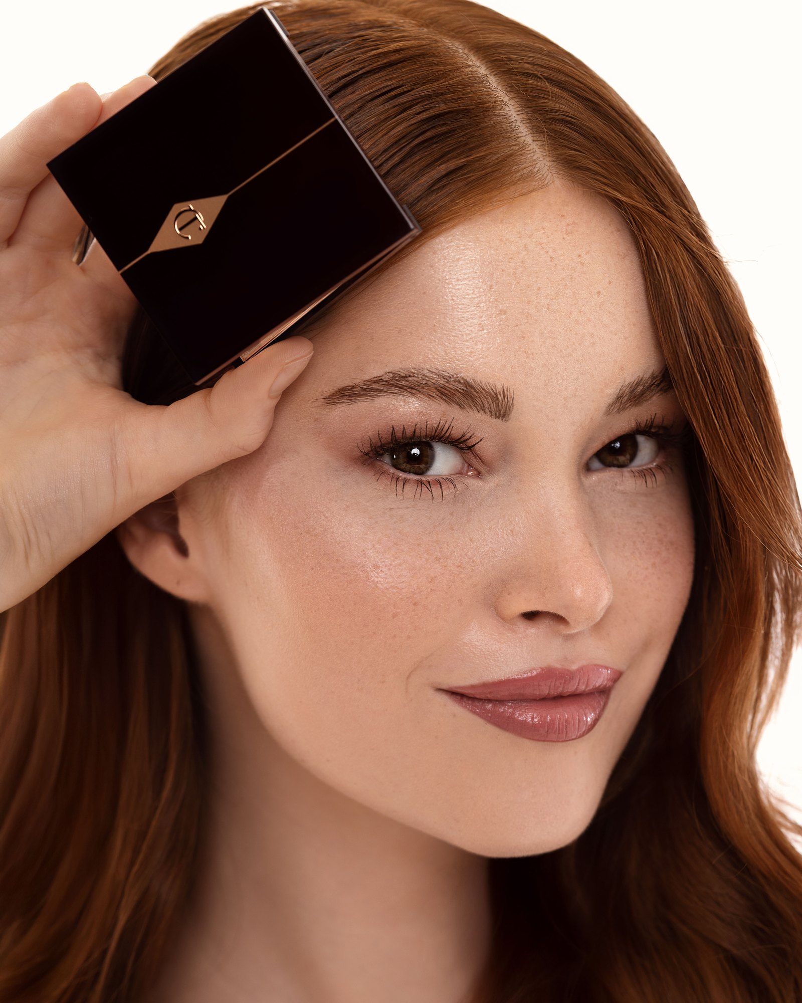 Woman with long auburn hair holding a black and rose gold Charlotte Tilbury compact against her head
