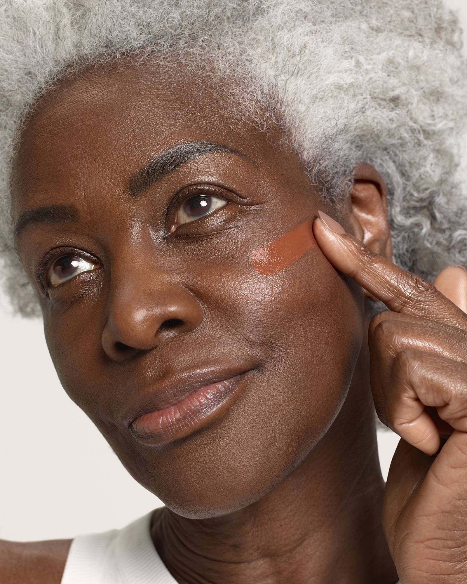 Older woman with silver curly hair smoothing a swatch of terracotta foundation onto her cheek