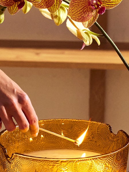 A hand lighting a large amber glass candle in a warm living room