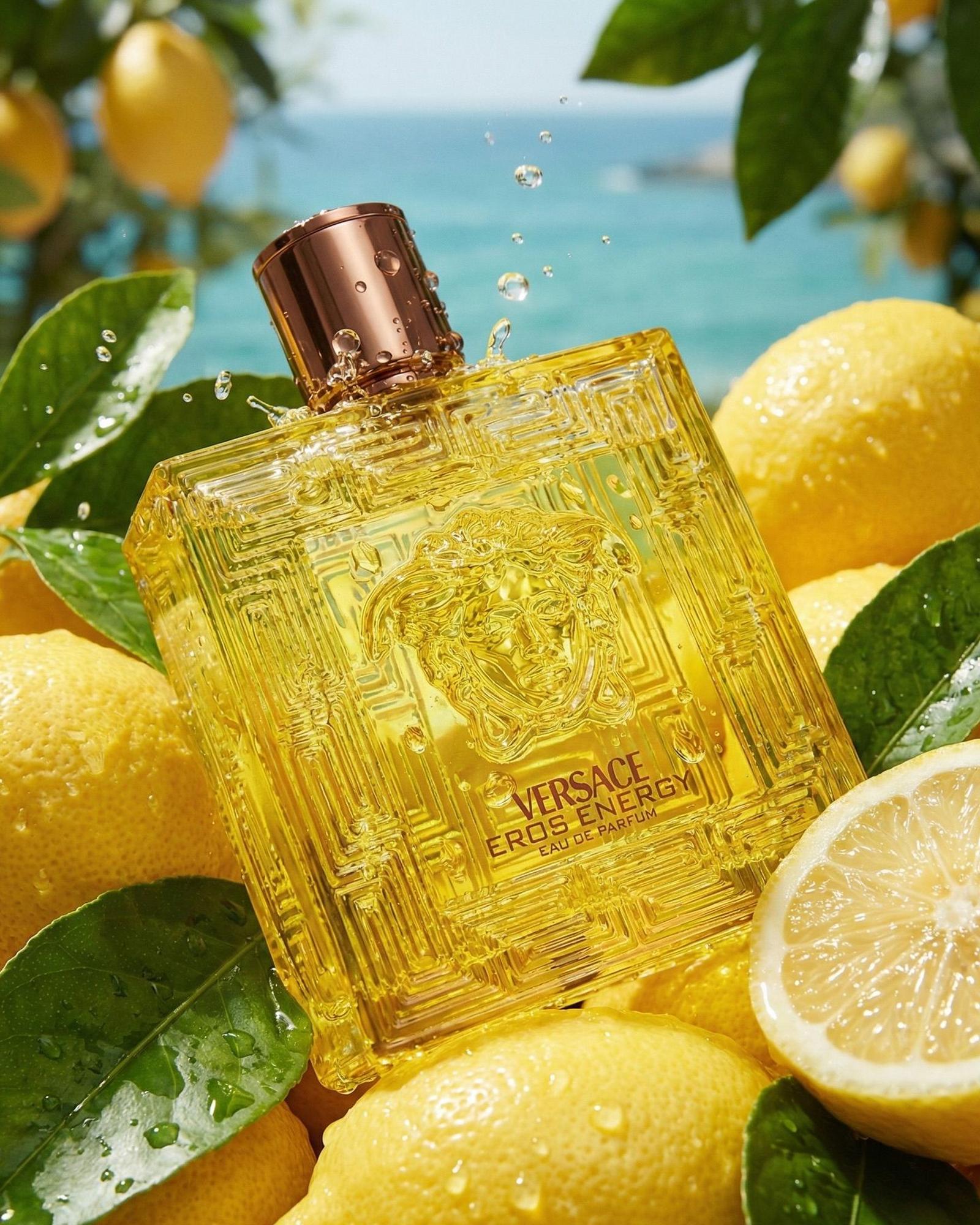 Yellow Versace Eros Energy bottle resting on wet lemons and leaves with a turquoise sea behind