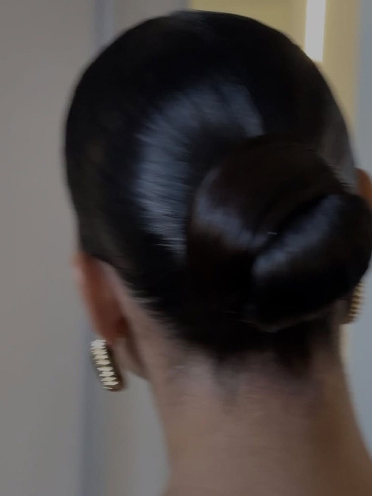 Back of a head with a glossy dark slicked-back low bun and gold ridged earrings, slightly blurred in motion