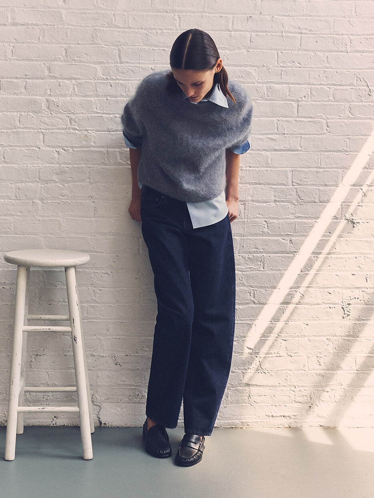 Rag & Bone model in a grey knit vest over a blue shirt and navy trousers by a white brick wall