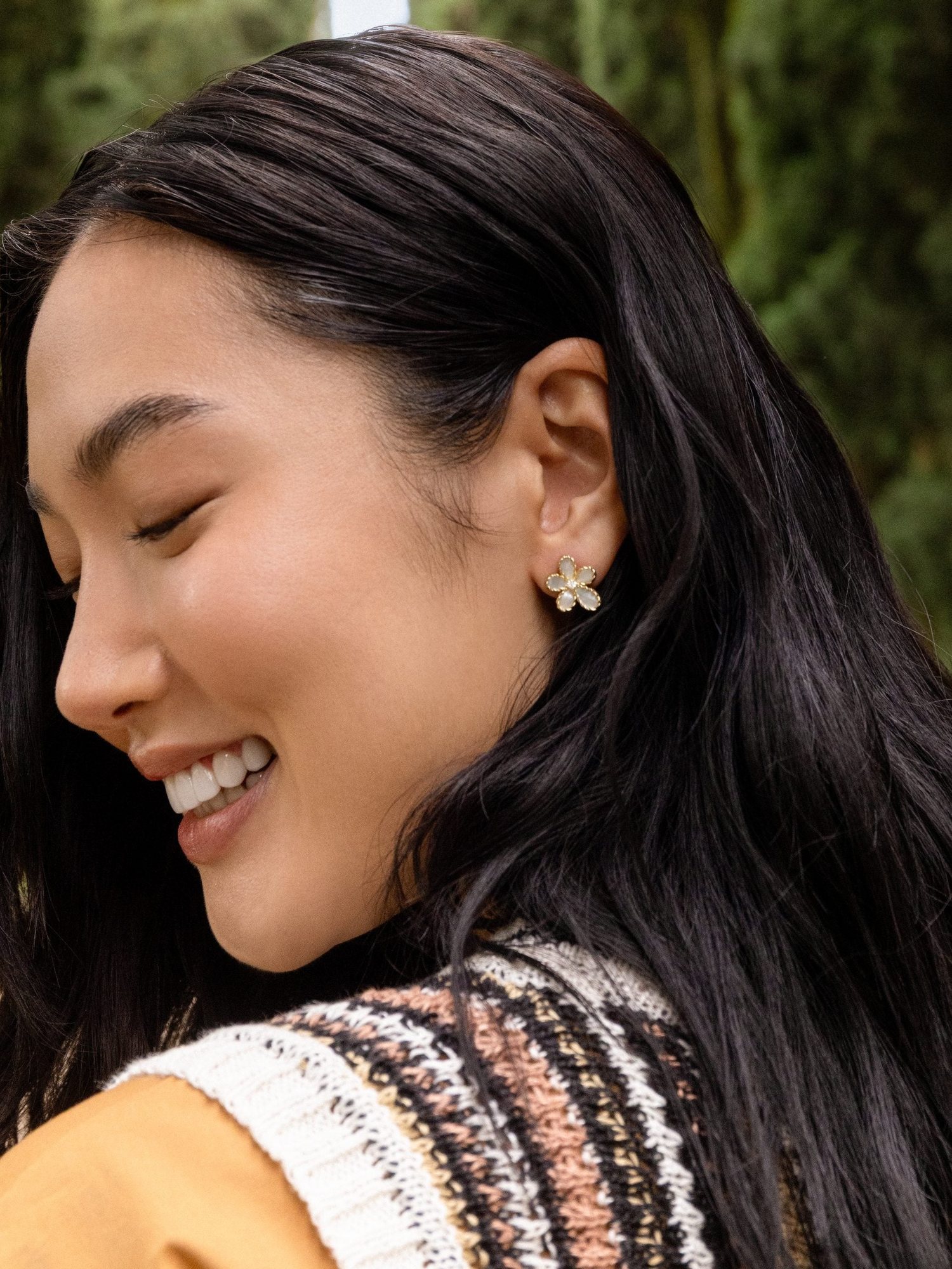 Smiling dark haired model in profile wearing flower stud earrings outdoors