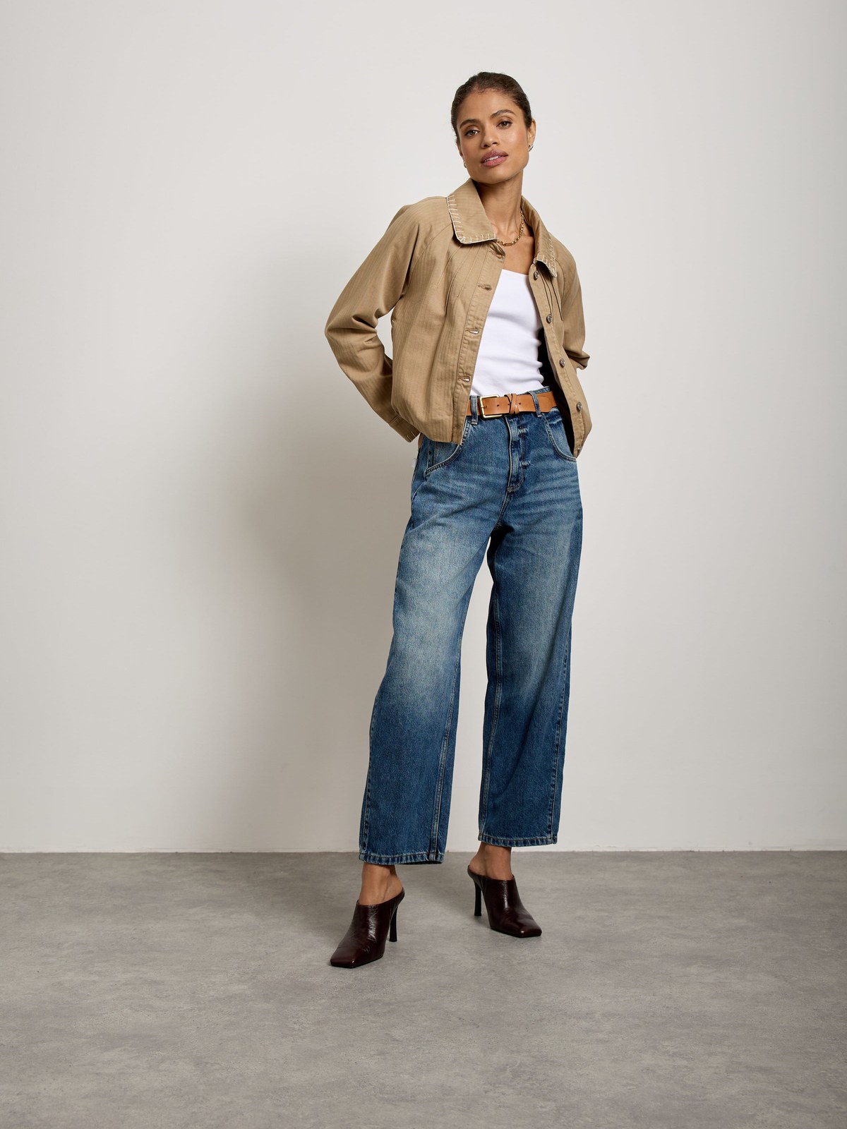 Model in a taupe jacket and blue wide-leg jeans on a pale studio set