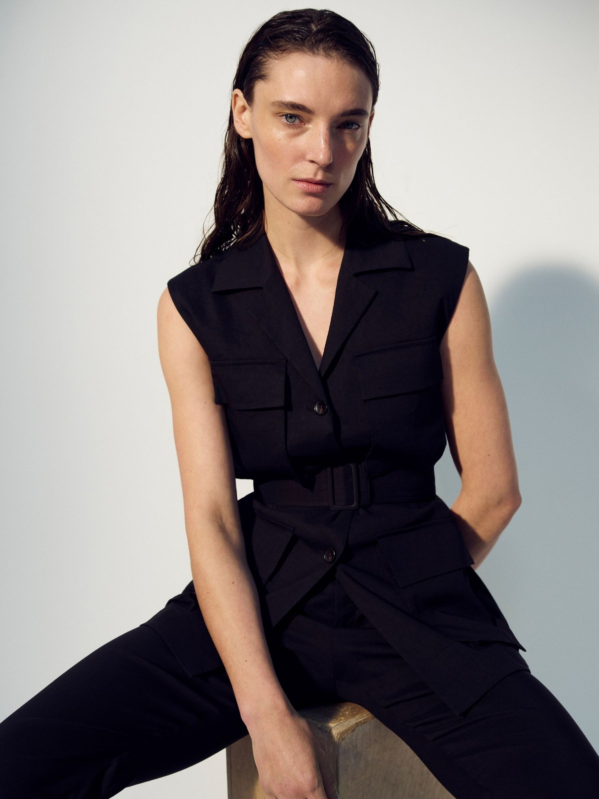 Model seated in a black sleeveless tailored jumpsuit against a pale studio backdrop