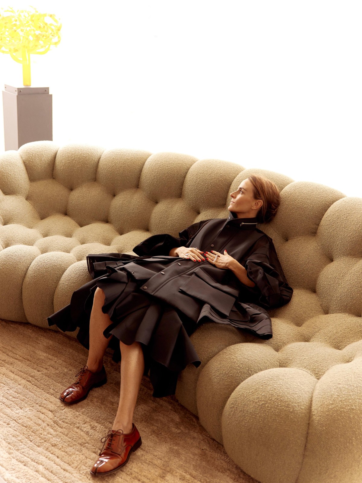 Woman reclining on a rounded beige sofa in a dark sculptural outfit
