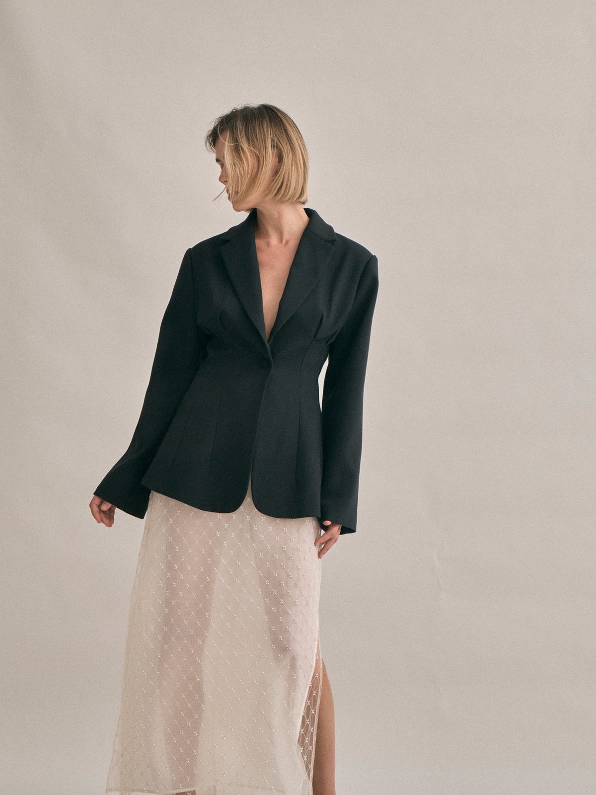Model in a black tailored blazer and ivory skirt against a studio wall