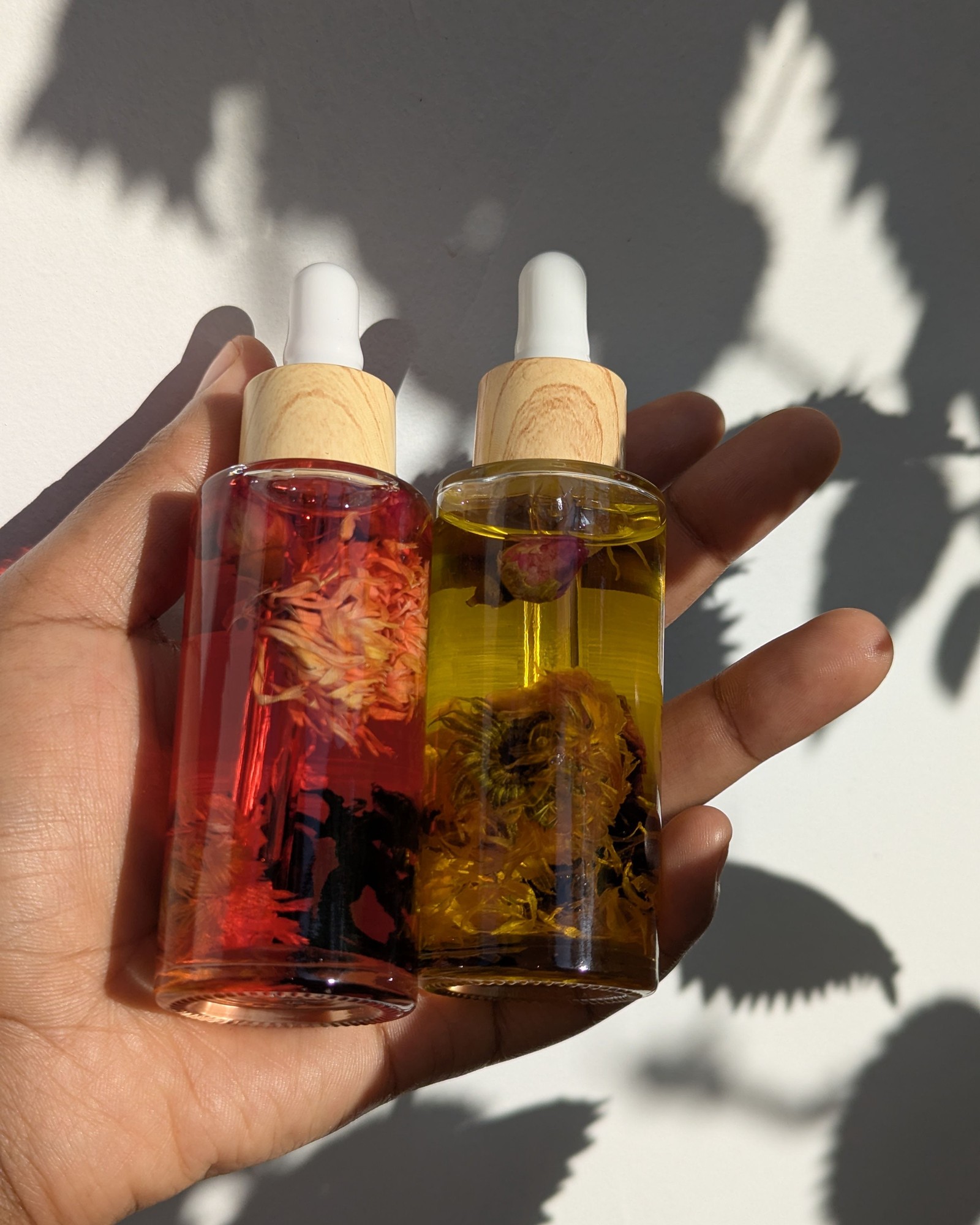 Hand holding two glass dropper bottles of oil filled with dried orange and yellow flowers in dappled sunlight
