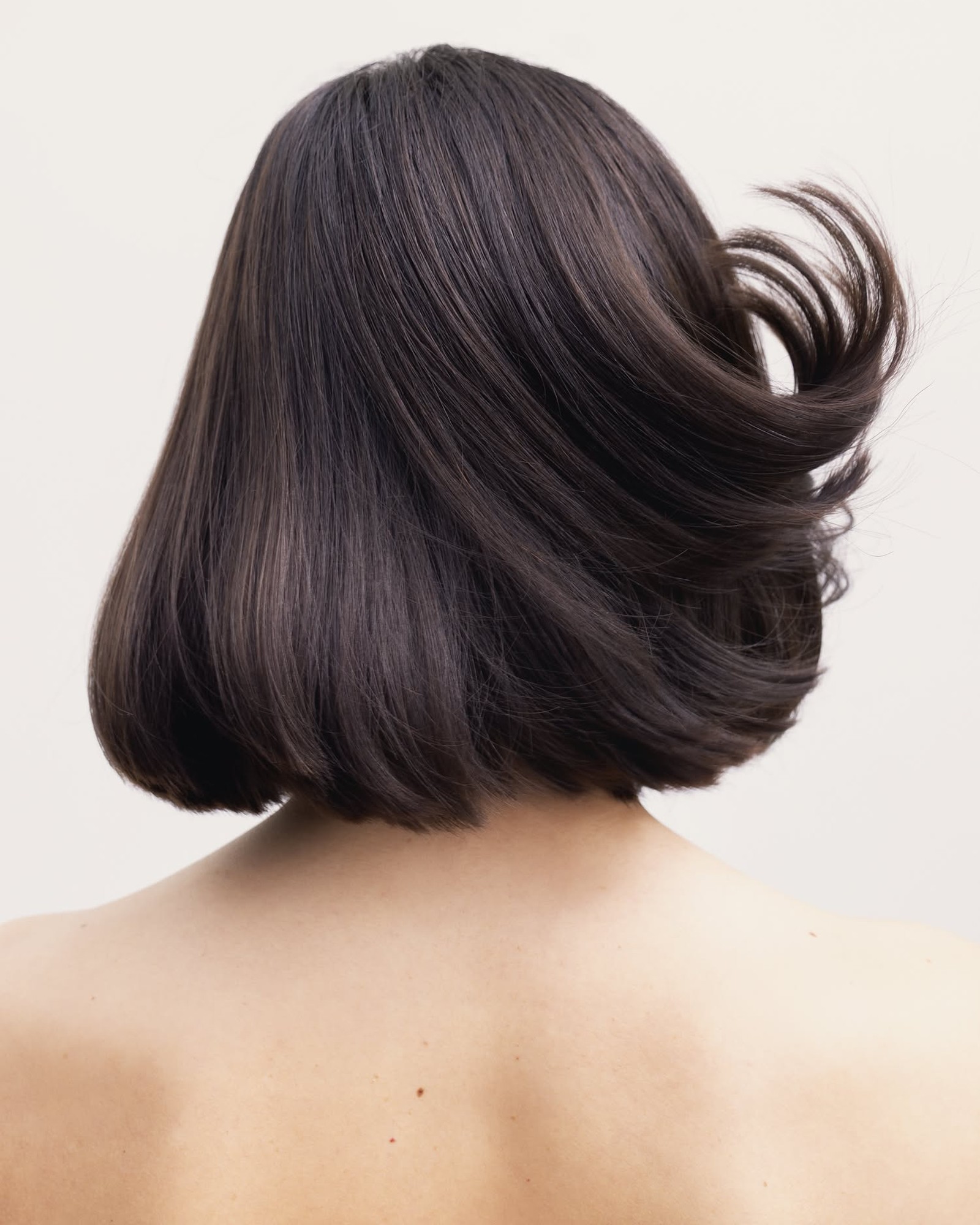 Back view of a woman with a glossy dark brown bob swinging against a pale wall