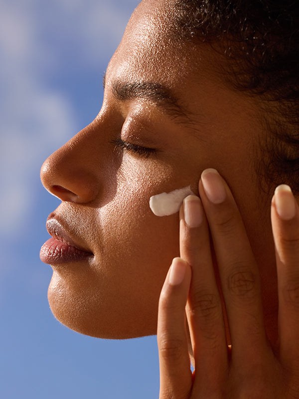 Woman applying sunscreen to her cheek in profile against a blue sky