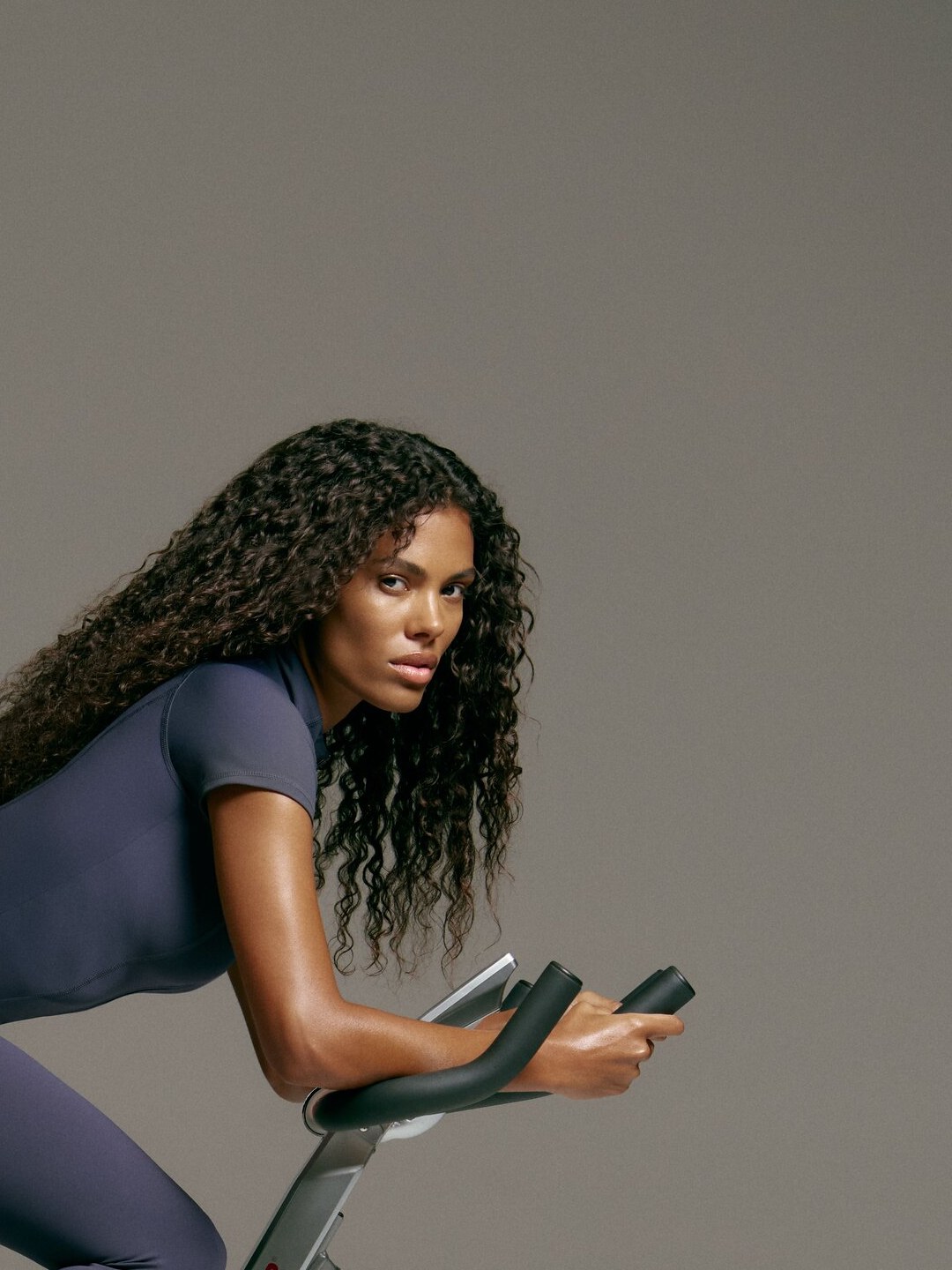 Tina Kunakey leaning over an exercise bike in a charcoal studio