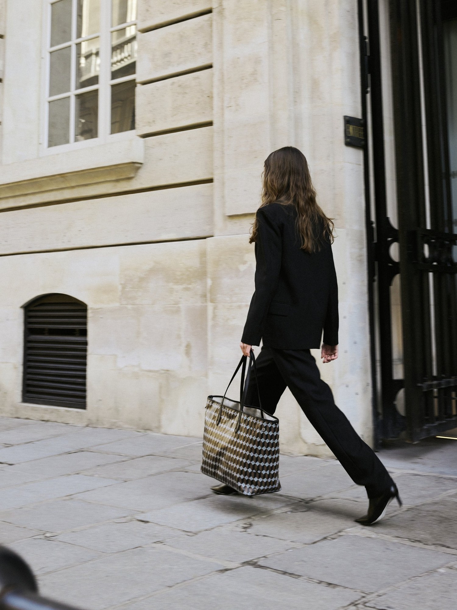 Woman in a black suit walking past a Paris stone building with a monogram tote