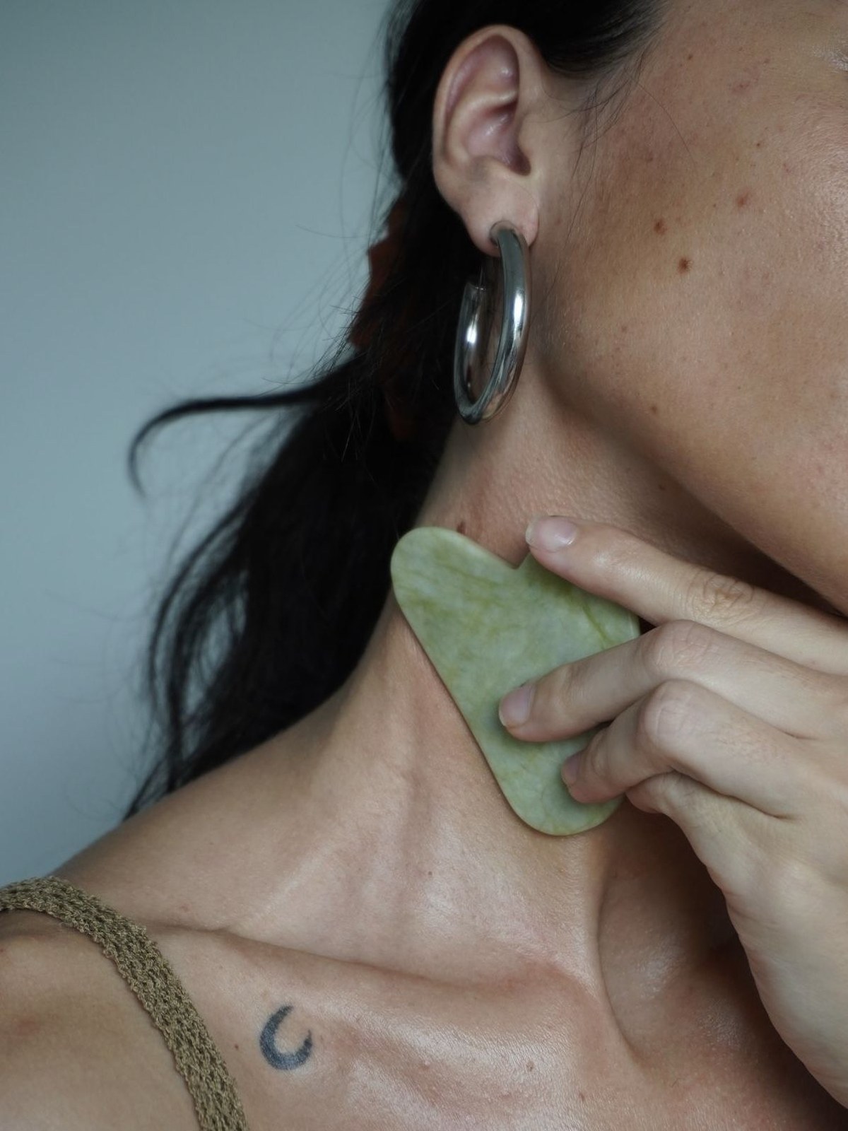 Woman in silver hoop earrings holding a heart-shaped green jade gua sha against her jaw and neck