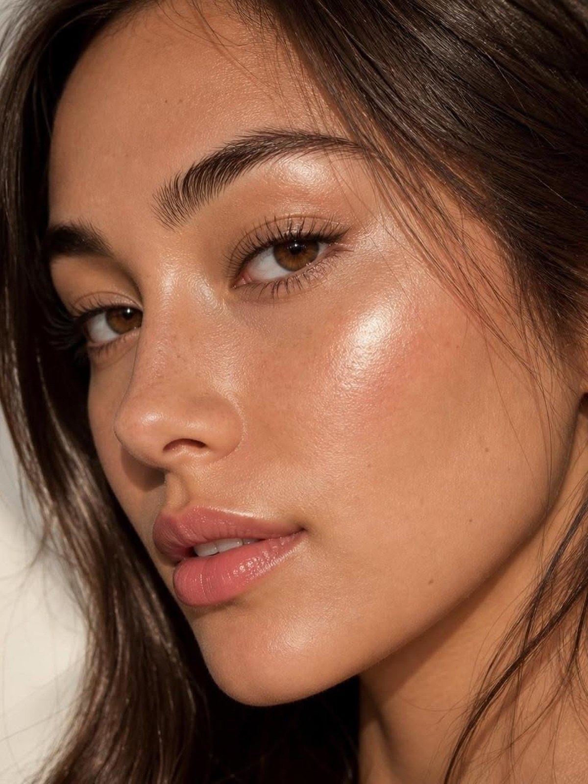 Close-up side profile of a woman with long brown hair and glowing tan skin with a bright highlight on the cheekbone