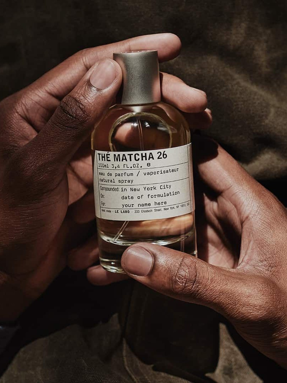 Two hands holding a bottle of Le Labo Thé Matcha 26 eau de parfum against a dark green jacket