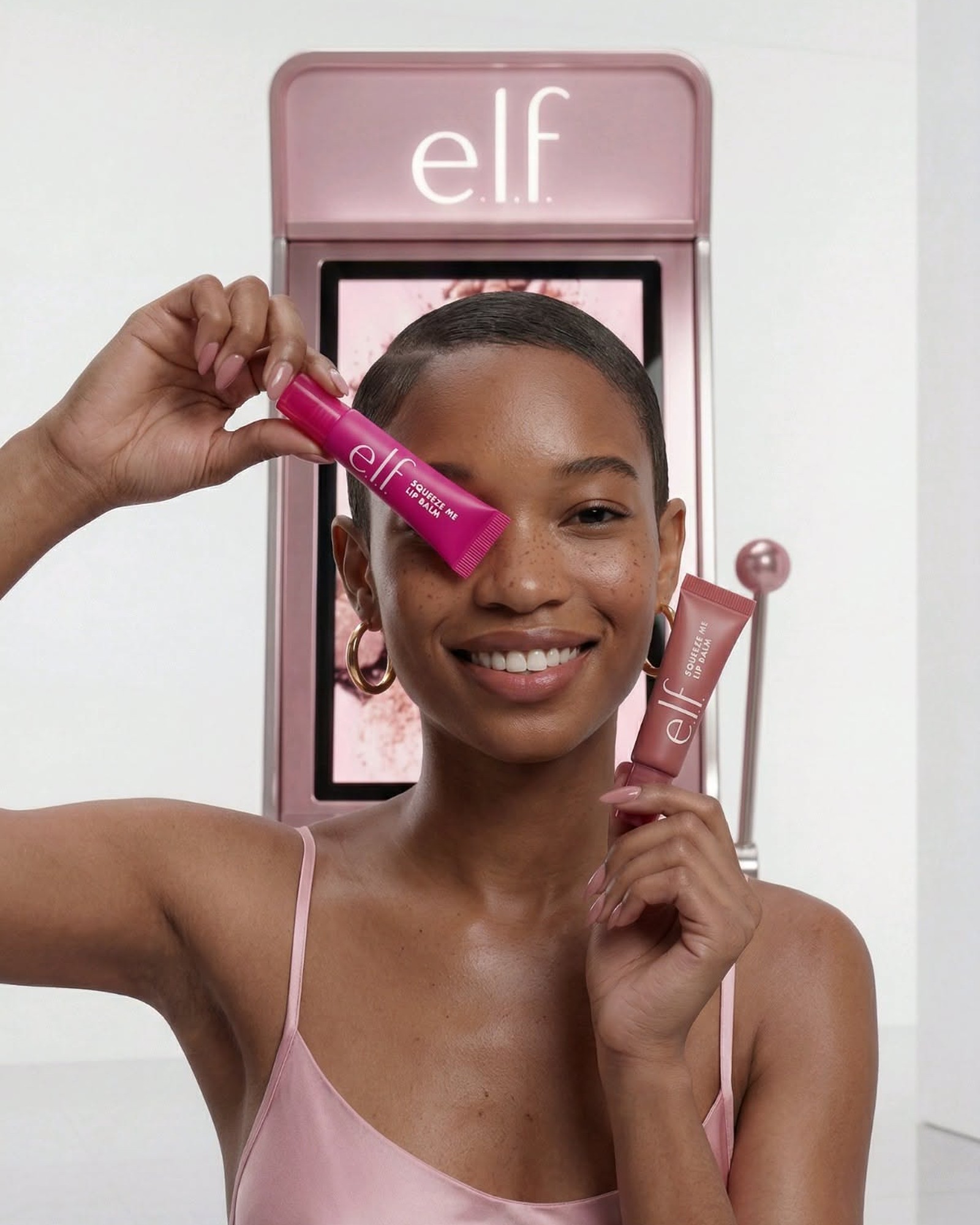 Smiling woman in a pink slip holding e.l.f. makeup in front of a pink e.l.f. booth