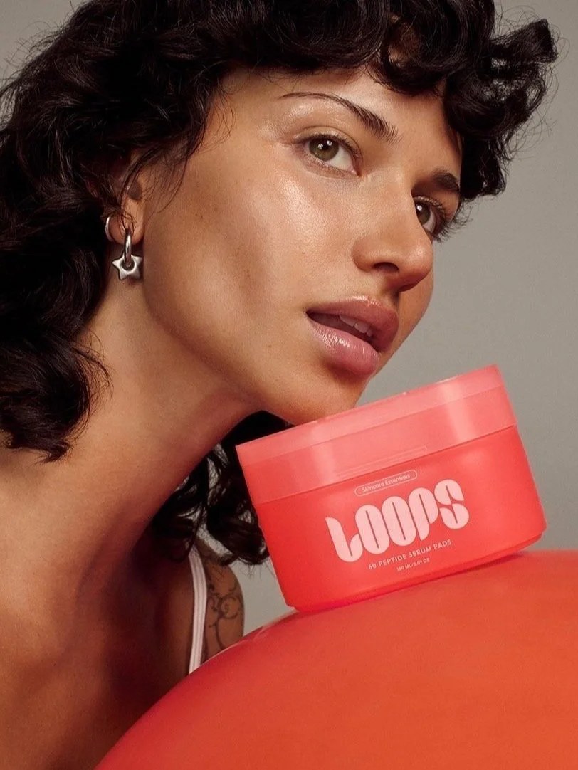 Curly-haired model holding a coral LOOPS skincare jar in a softly lit studio