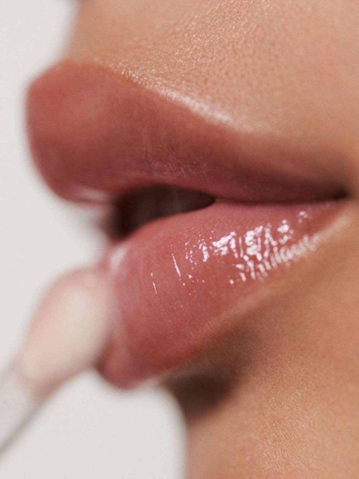 Side profile of shiny rose lips with a gloss wand blurred at the edge of the frame