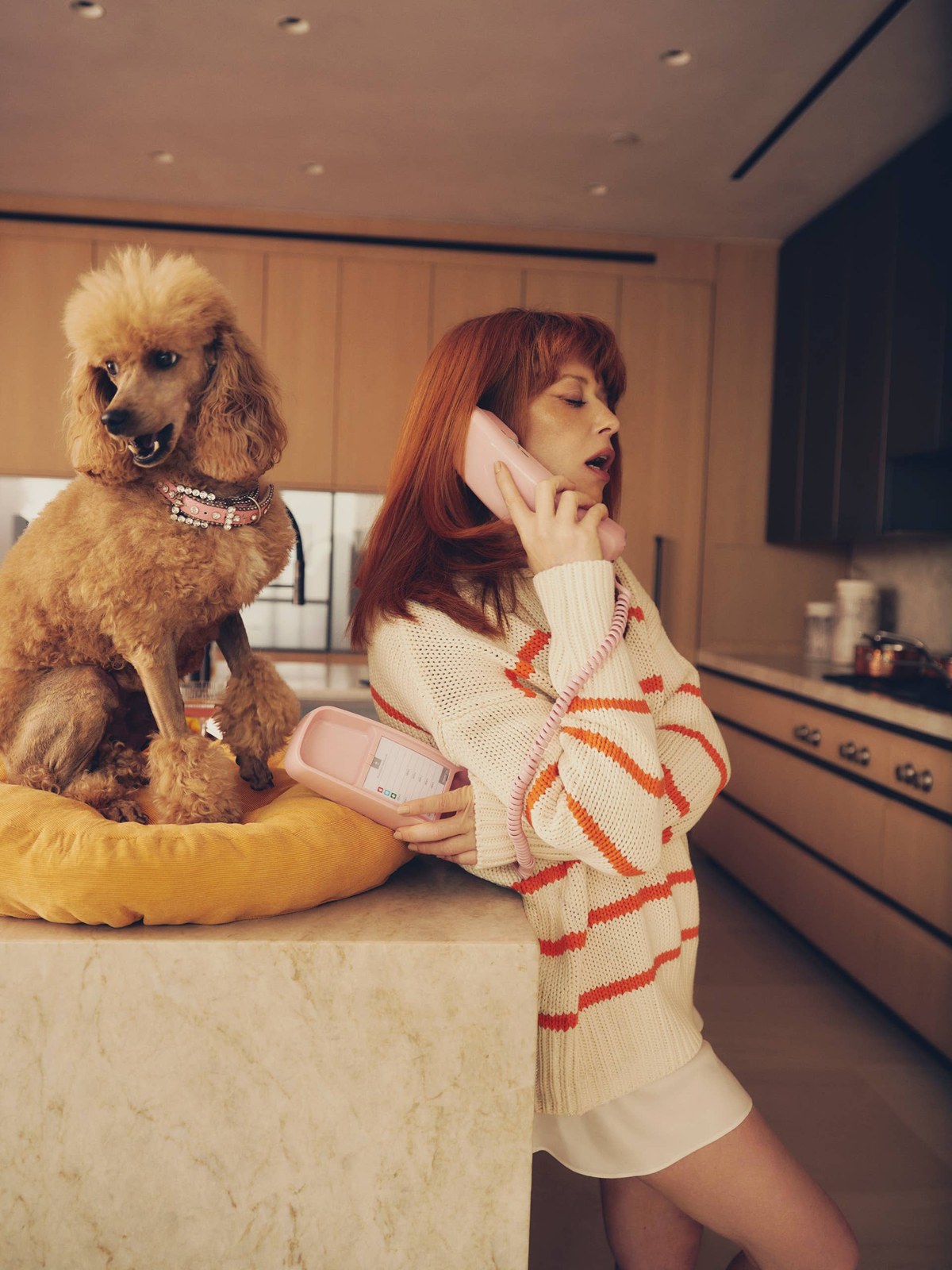 Natasha Lyonne in a cream and orange striped sweater in a kitchen with a poodle