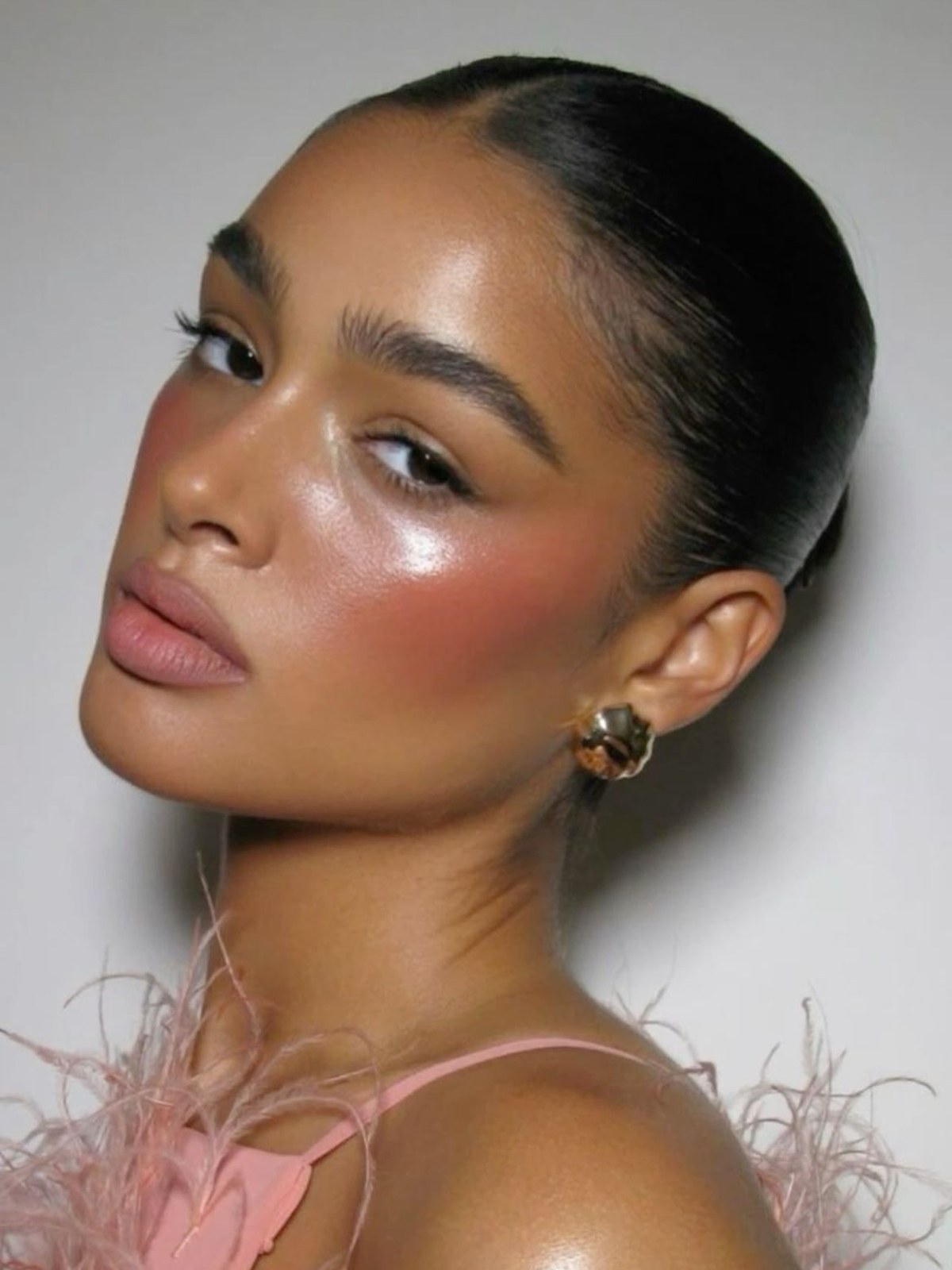 Woman with a sleek dark bun, glowing skin, pink cheek color and a pink feathered top