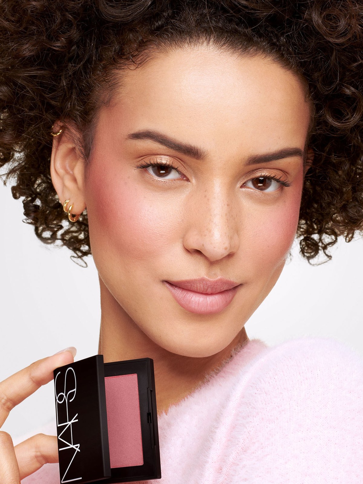 Curly haired model in a pink sweater holding a mauve NARS blush compact