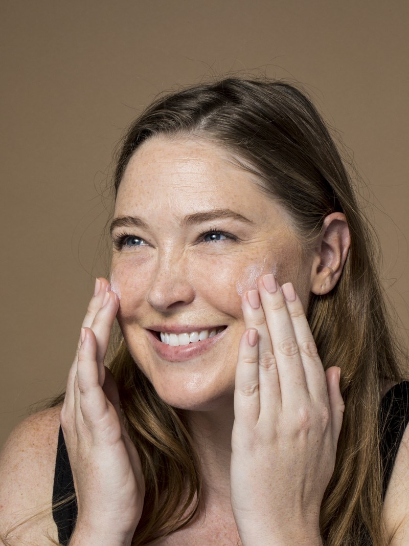 Model smiles while applying moisturizer to both cheeks in a warm studio image.