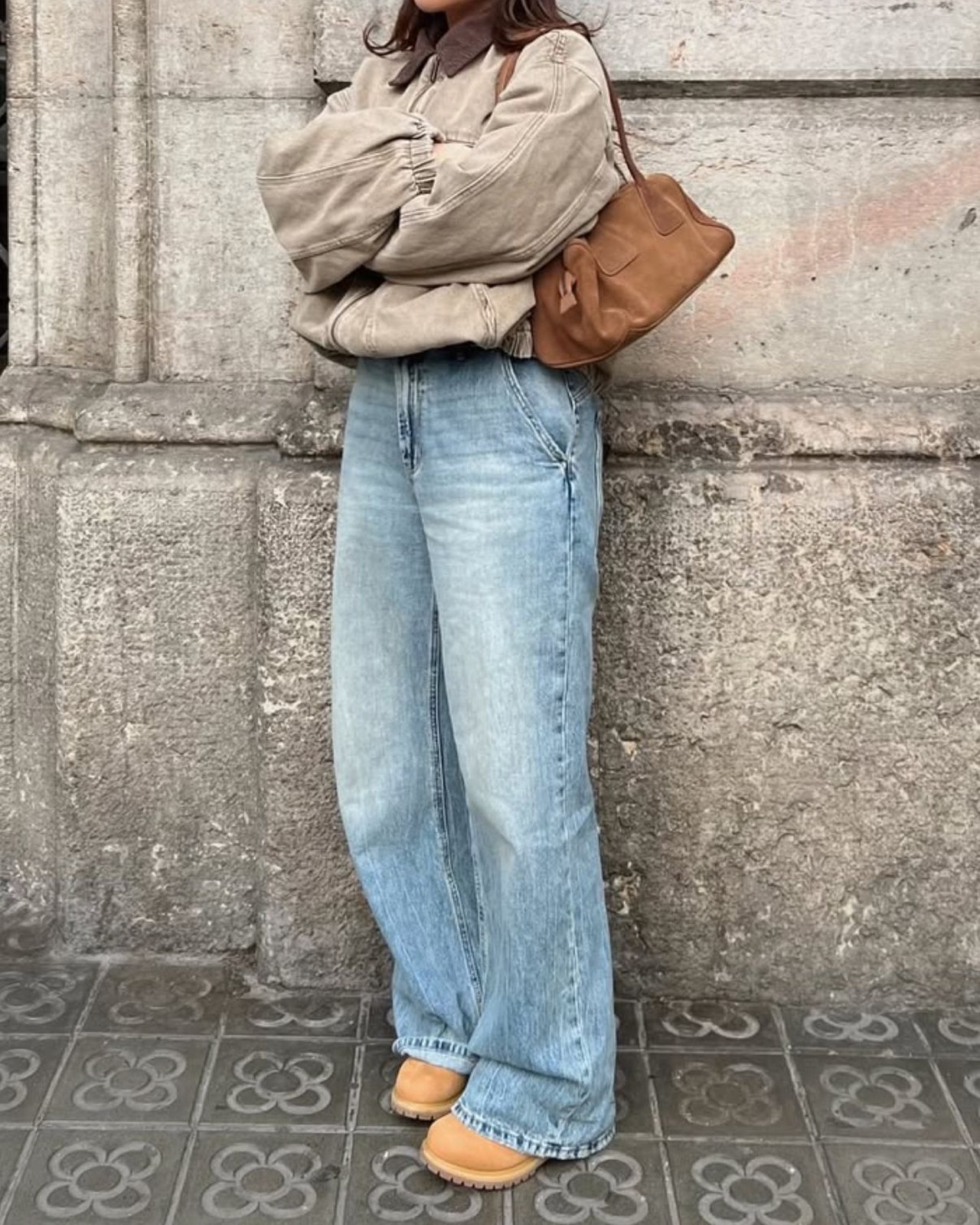 Woman against a stone wall in a beige bomber jacket, baggy light jeans and wheat Timberland boots with a tan bag