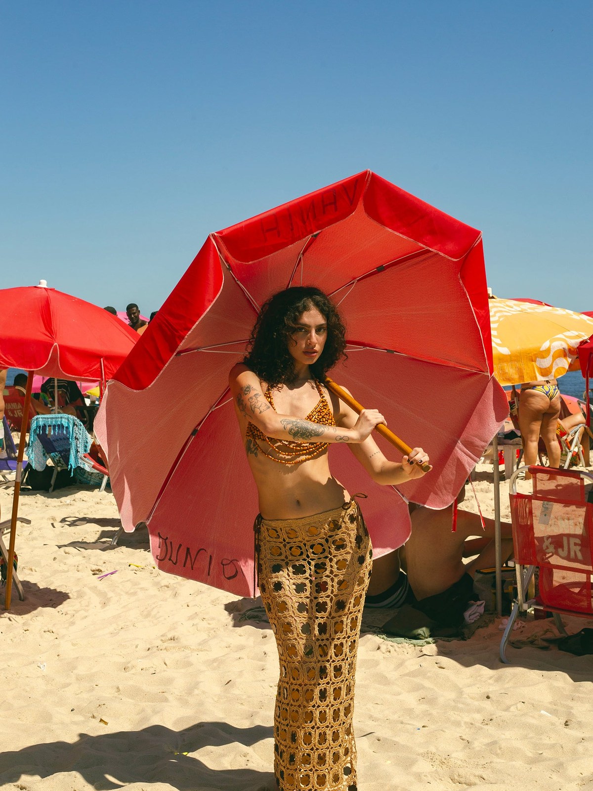 Woman in a crochet beach outfit beneath a red parasol by the shoreline