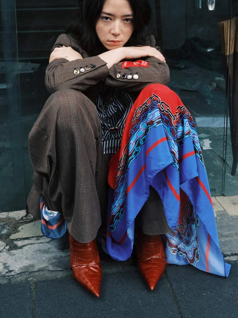 Model in a grey outfit with a blue and red drape seated by a city storefront