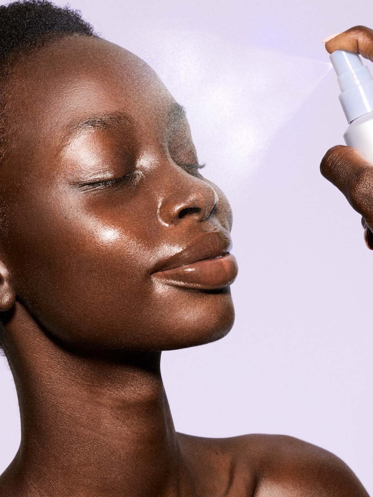 Profile of a model with glowing skin being spritzed with face mist on a lavender background