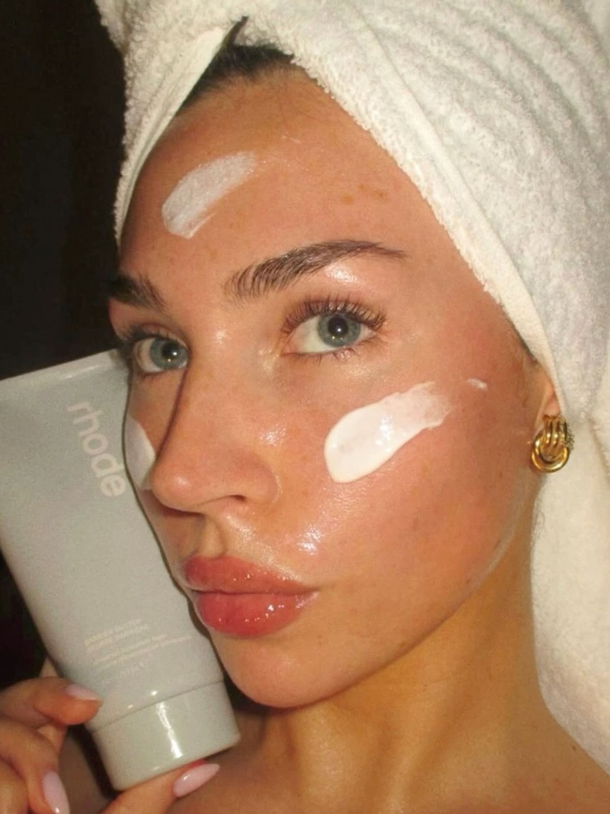 Woman in a towel turban applying rhode moisturizer to her cheek