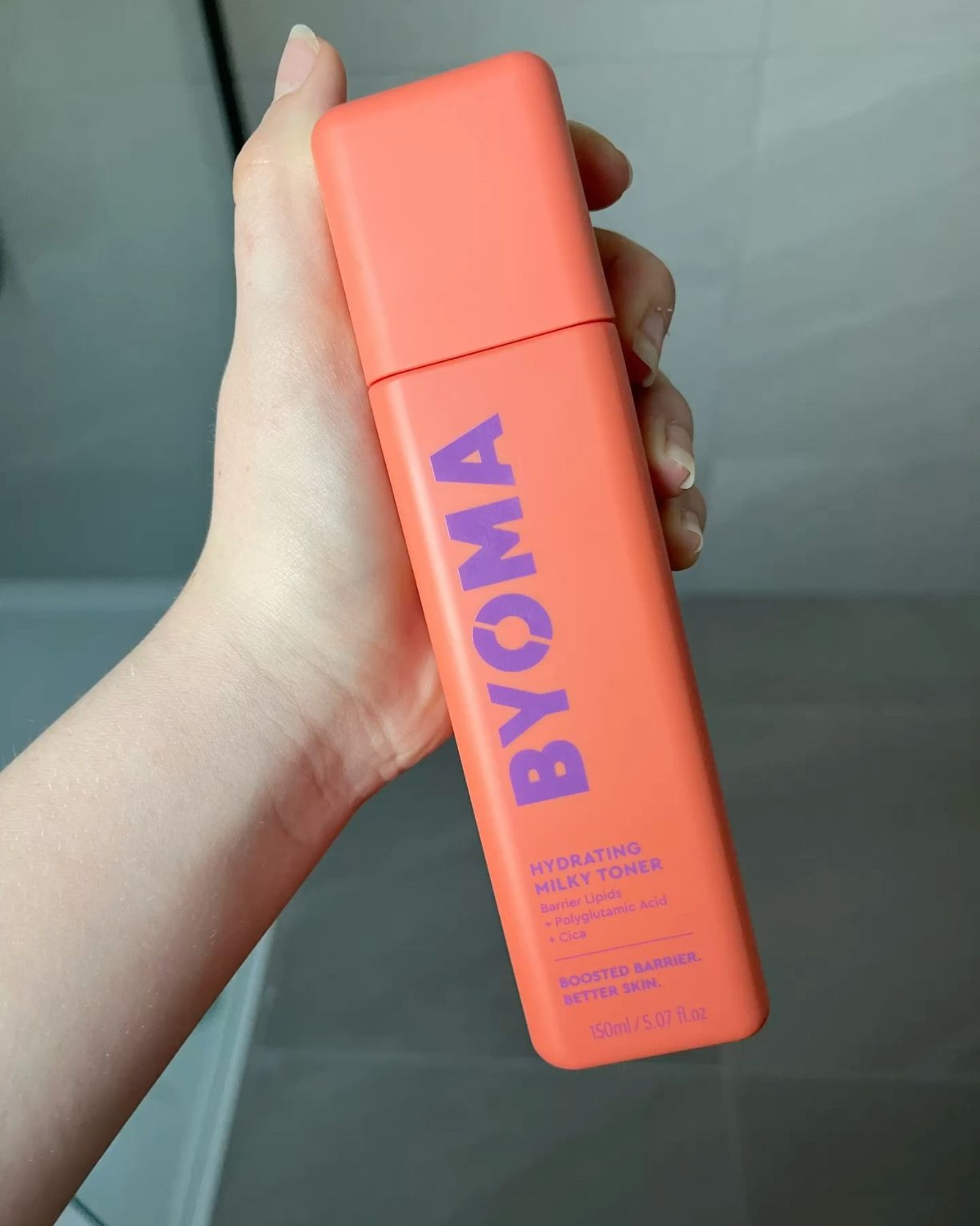 Hand holding a coral BYOMA Hydrating Milky Toner bottle in front of grey bathroom tiles