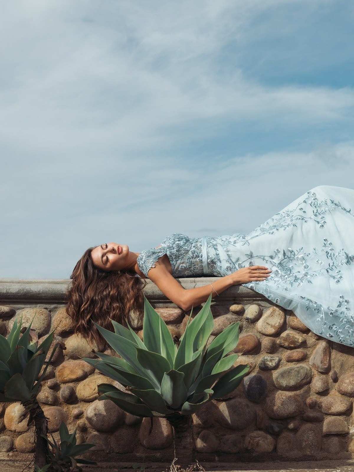 Model reclining on a stone wall among agave in a powder blue embroidered tulle dress