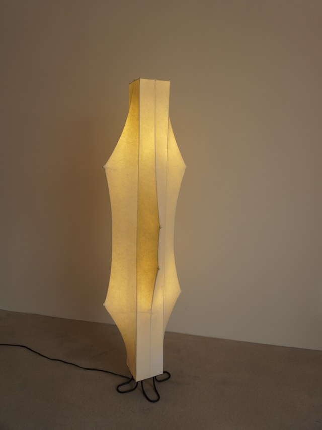 Warmly lit sculptural floor lamp against a taupe studio wall