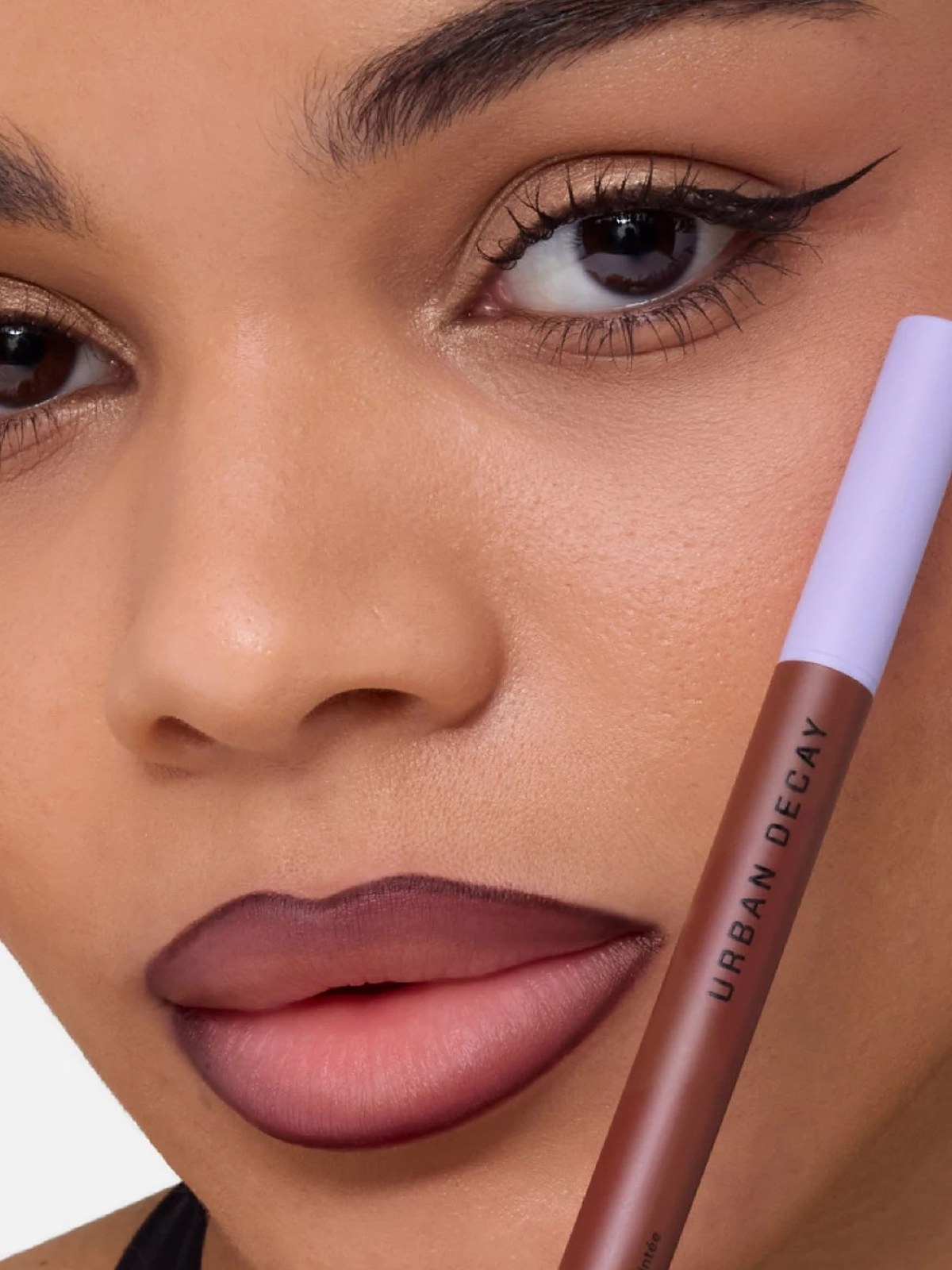 Model with winged liner holding a lilac lip pencil to a mauve lip
