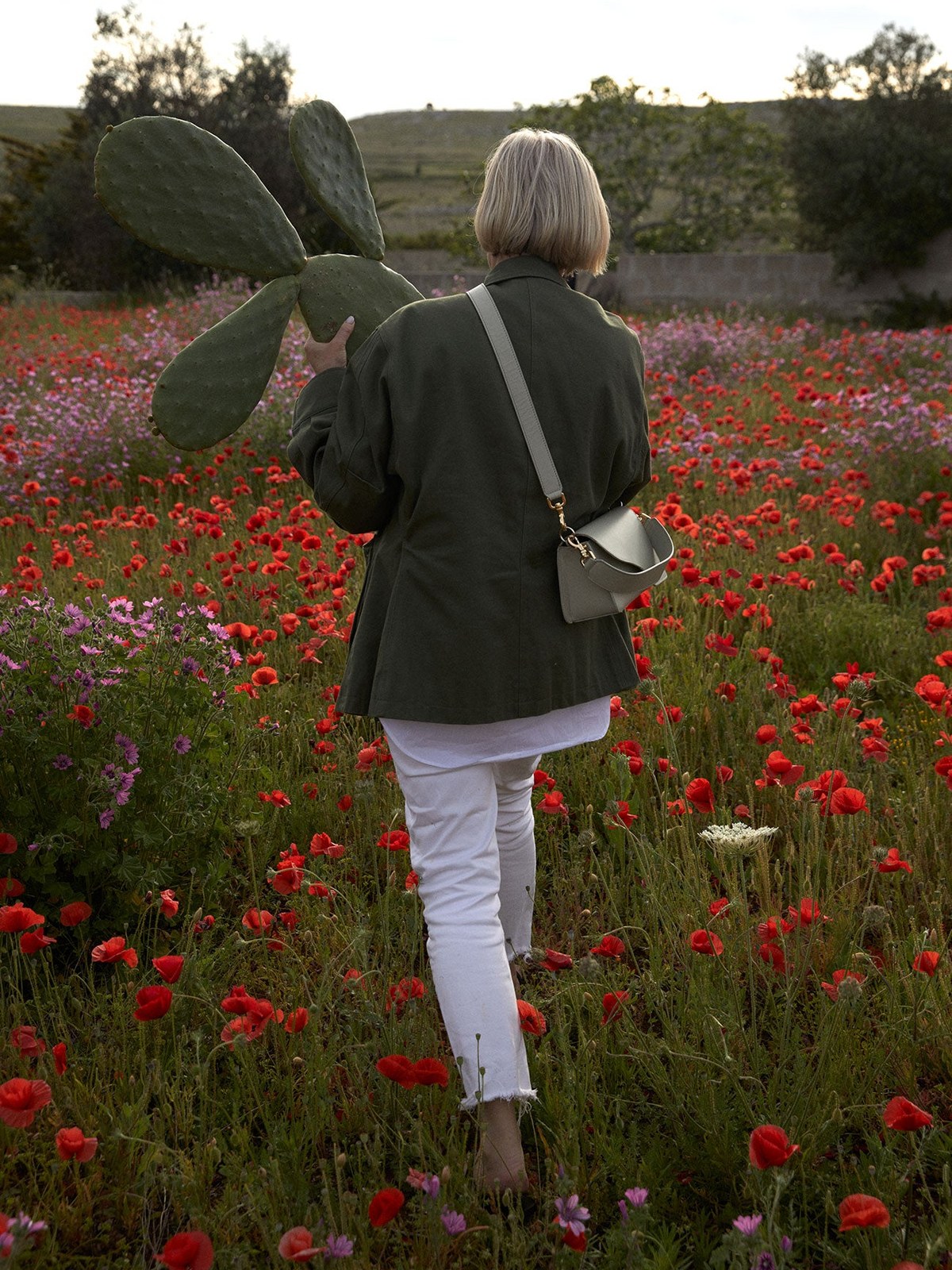 Woman seen from behind in a flowered field carrying a small shoulder bag.